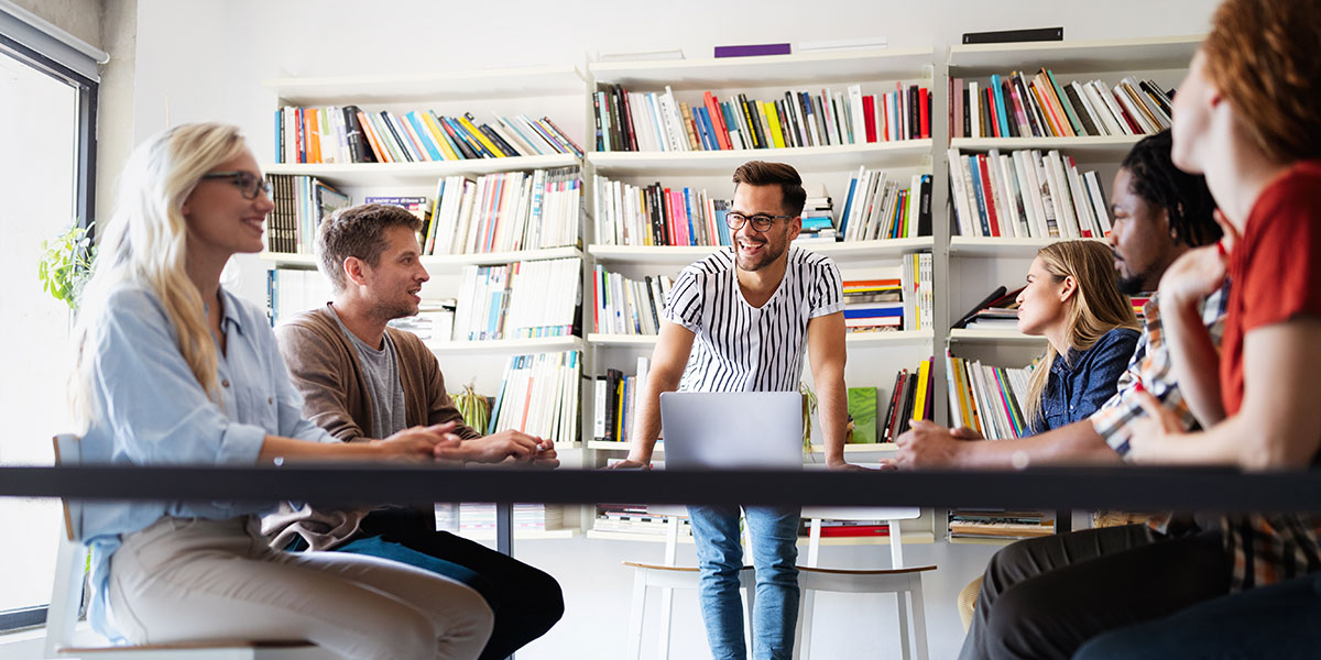 A UX designer stands at a desk leading five seated designers in the ideation design process 