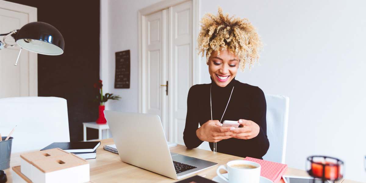 An aspiring UX designer sitting at a desk with a laptop and cup of coffee, working on her phone