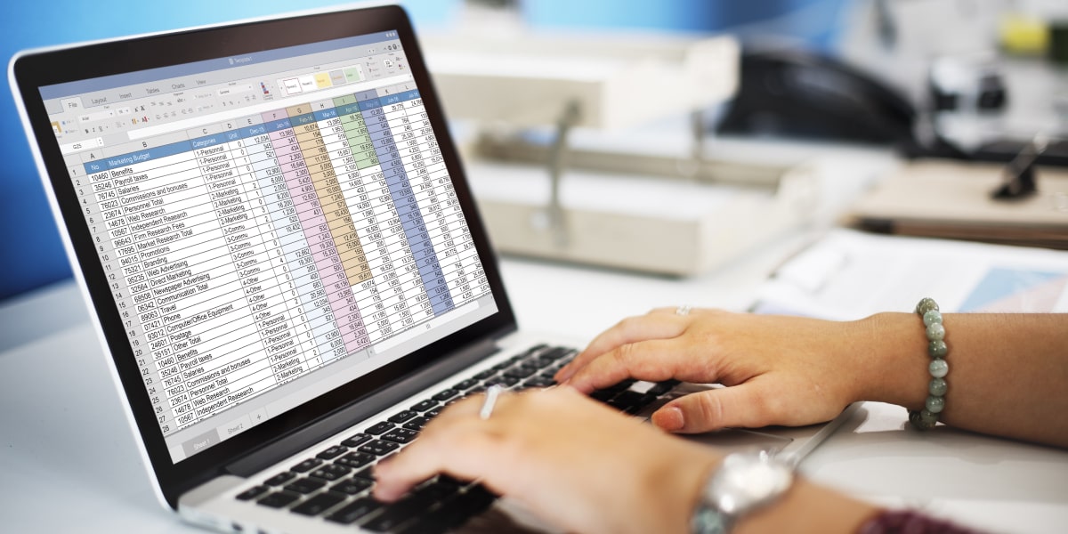 A pair of hands typing on a keyboard, with a spreadsheet of data on a laptop screen